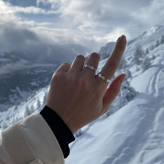 Zu sehen ist eine Hand mit zwei Ringen. Dem Antiwave Big und dem Smileyring. Im Hintergrund sind verschneite Berge.