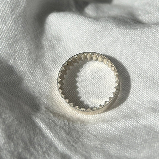 Ein Foto vom Tastsinntraining-Ring "TIA 02 Riffel" auf einem weißen, Leinenuntergrund. Der Ring hat einen glatten Look von außen und kleine Zacken innen.